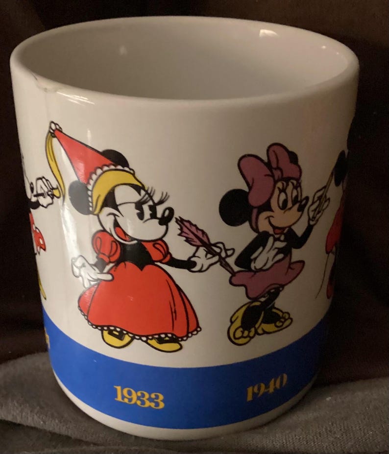 May include: White ceramic mug featuring Minnie Mouse in various outfits. The mug has a blue band with the years 1933 and 1940 printed on it. The mug has a few small chips on the rim.