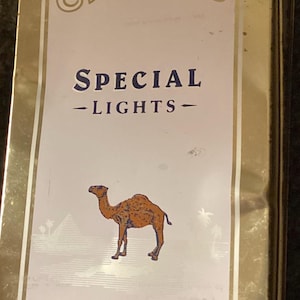 May include: Vintage Camel Special Lights cigarette tin. The tin has a gold border and a white background with the Camel logo at the top. The text "SPECIAL - LIGHTS -" is in the center, with a camel illustration below. "Special Mild Blend" is at the bottom.