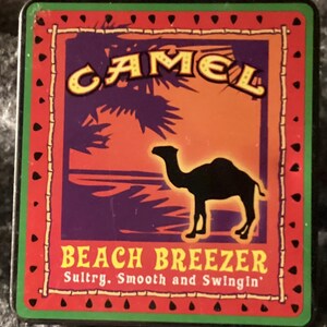 May include: Rectangular metal sign with a red border and a green edge. The sign features the word "CAMEL" in yellow letters, a black camel silhouette, and the words "BEACH BREEZER" in yellow. The background has purple, orange, and palm tree graphics.