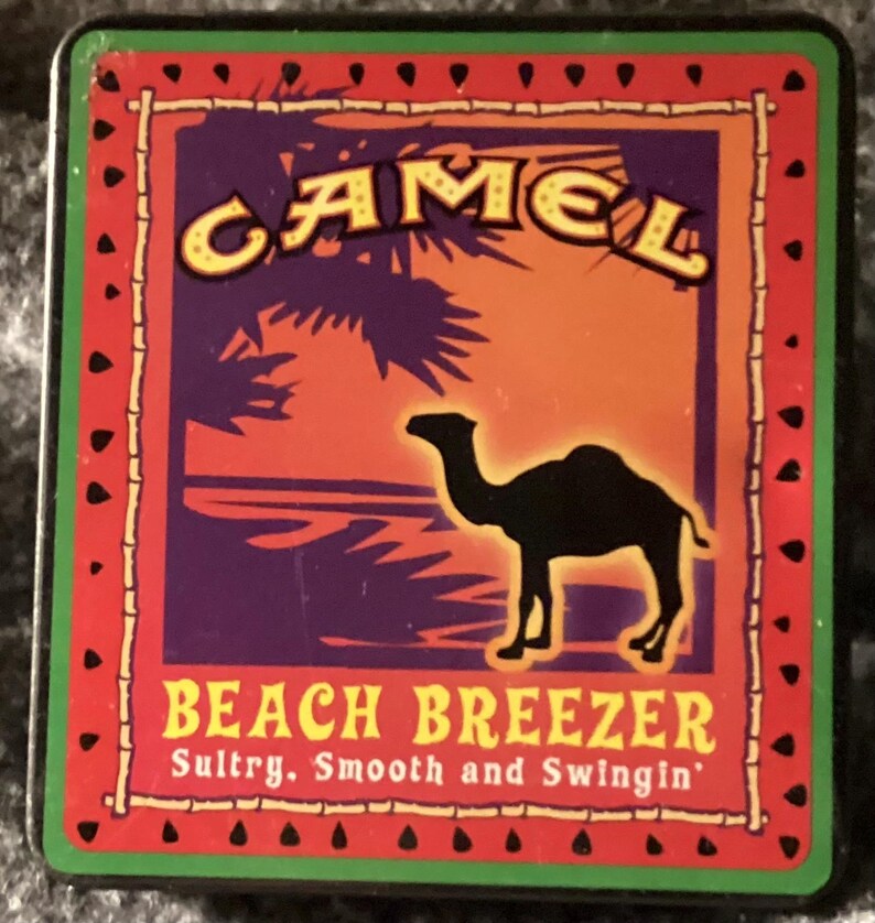 May include: Rectangular metal sign with a red border and a green edge. The sign features the word "CAMEL" in yellow letters, a black camel silhouette, and the words "BEACH BREEZER" in yellow. The background has purple, orange, and palm tree graphics.