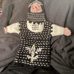 May include: Handmade felt puppet of a medieval knight. The puppet has a black and white patterned tunic, gray sleeves, and a helmet with a smiling face. It holds a black felt sword. A unique, handcrafted toy.
