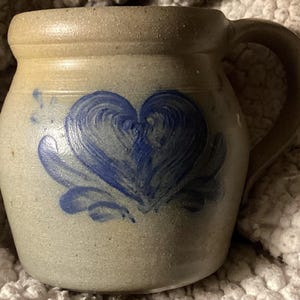 May include: A beige stoneware mug with a large blue heart design. The mug has a rounded body, a sturdy handle, and a textured surface. The heart is painted with brushstrokes, adding a rustic charm to the handmade pottery.