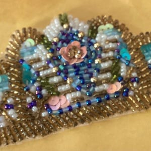 May include: A beaded applique in a cloud shape, featuring gold, blue, pink, and white beads. The design includes a central flower and accents of sequins and seed beads, creating a textured, decorative embellishment.