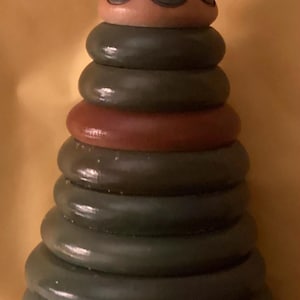 May include: A wooden stacking toy featuring a painted face on top, a blue bonnet, and a series of green and red rings. The base is decorated with a scalloped design. A classic toy for children.