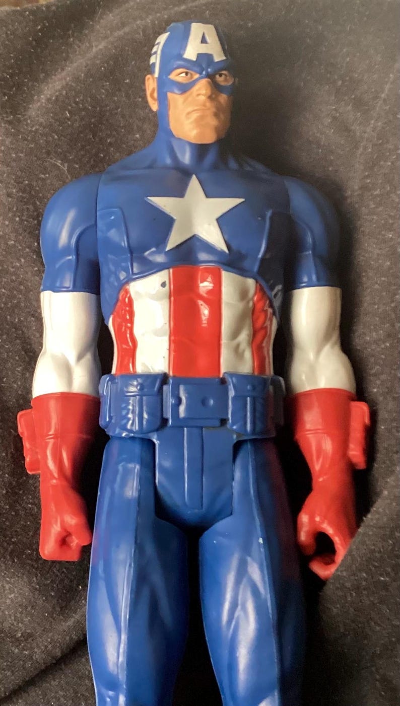 May include: A Captain America action figure. The figure is predominantly blue, red, and white, with a large white star on the chest. The head is blue with a white "A" on the forehead. The figure has red gloves and boots.
