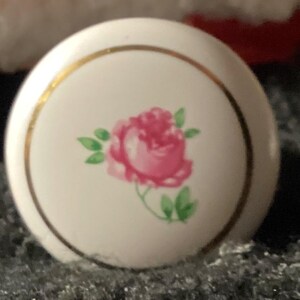 May include: A round, white cabinet knob with a gold rim. The center features a pink rose with green leaves. This decorative knob is suitable for furniture and adds a touch of floral elegance.
