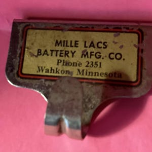 May include: Vintage metal battery clip with a rectangular label. The label has the text "MILLE LACS BATTERY MFG. CO. Phone 2351 Wahkon Minnesota" in black font on a yellow background with a red and black border. The clip has a curved hook.