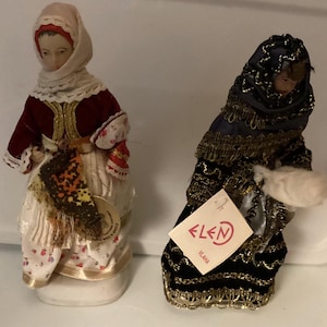 May include: Two vintage dolls in traditional attire. The doll on the left wears a red velvet jacket, white dress with floral print, and a headscarf. The doll on the right is dressed in a dark blue outfit with gold trim and a head covering. Both dolls are on white bases.