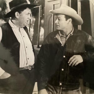May include: Black and white photograph of two men in cowboy attire. One man wears a black hat, vest, and button-down shirt. The other man wears a cowboy hat, jacket, and button-down shirt. The background is a blurred outdoor setting.