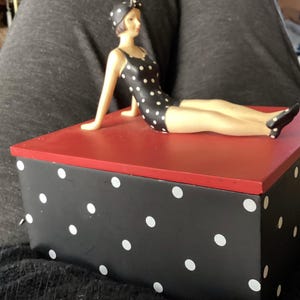 May include: A decorative box with a red top and black sides adorned with white polka dots. A figurine of a woman in a black and white polka dot swimsuit and matching cap is resting on the box. The figurine is in a reclining pose.