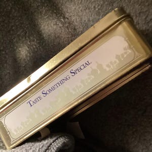 May include: A rectangular, gold-colored tin with the words "TASTE SOMETHING SPECIAL" in blue lettering. The tin has a white label with a decorative border and a silhouette design. The tin is slightly open, revealing its contents.