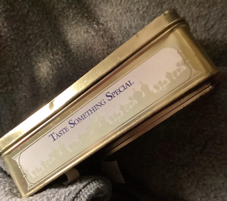 May include: A rectangular, gold-colored tin with the words "TASTE SOMETHING SPECIAL" in blue lettering. The tin has a white label with a decorative border and a silhouette design. The tin is slightly open, revealing its contents.