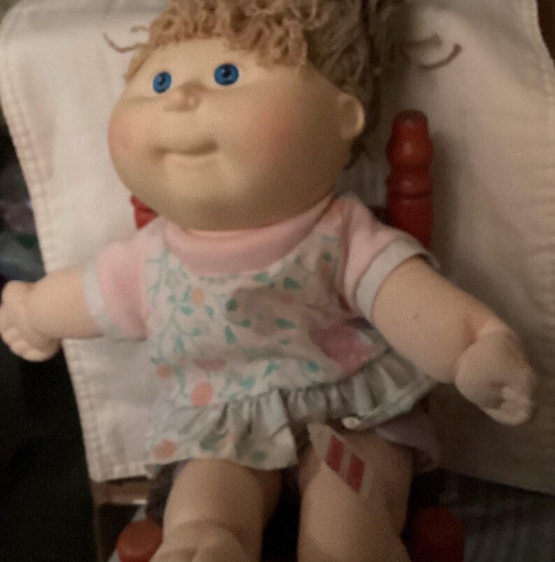 May include: A vintage Cabbage Patch Kid doll with blue eyes and light brown curly hair. The doll is wearing a pink and white floral dress with a ruffled skirt and a pink shirt. The doll is sitting in a red wooden chair.