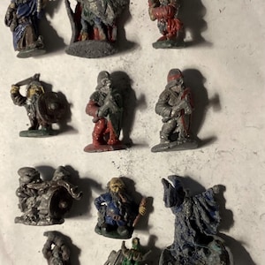 May include: A collection of miniature fantasy figurines, hand-painted in various colors. The figures depict warriors, wizards, and other characters, with details like shields, weapons, and robes. The figurines are on a neutral surface.