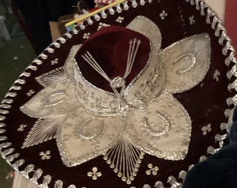 May include: A burgundy sombrero with intricate silver embroidery and embellishments. The hat features a wide brim with a floral design and a velvet crown. The brim is trimmed with silver braid.