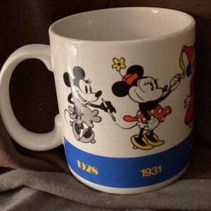 May include: White ceramic mug featuring Minnie Mouse illustrations in black, red, and yellow. The mug has a blue band with the years 1928 and 1931 in gold. The handle is white and the interior is also white.