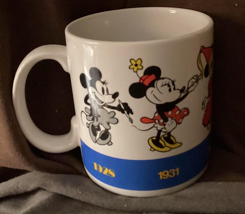 May include: White ceramic mug featuring Minnie Mouse illustrations in black, red, and yellow. The mug has a blue band with the years 1928 and 1931 in gold. The handle is white and the interior is also white.