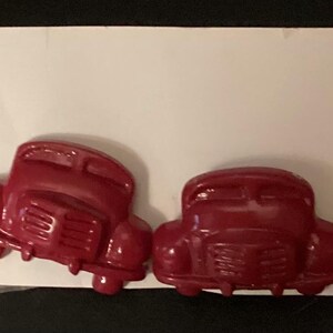 May include: Four small, red vintage car-shaped embellishments are arranged in a row. The cars have detailed features, including fenders, headlights, and a trunk. The embellishments are likely for crafts or scrapbooking.