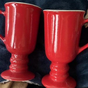 May include: Two red ceramic mugs with a pedestal base and a curved handle. The mugs have a glossy finish and a white interior rim. The mugs are designed for hot beverages.