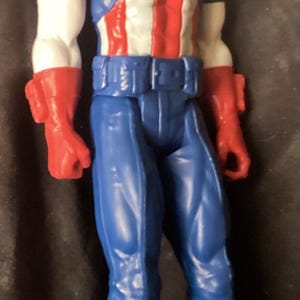 May include: A Captain America action figure. The figure is wearing a blue and white helmet, a blue suit with a white star, and a red, white, and blue torso. The figure has red gloves and boots.