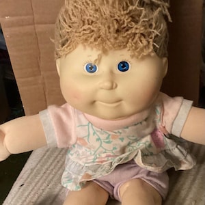 May include: A vintage doll with a soft, fabric body and a vinyl head. The doll has light skin, blue eyes, and light brown yarn hair. It wears a pink and floral print top and lilac shorts. The doll's arms and legs are extended.