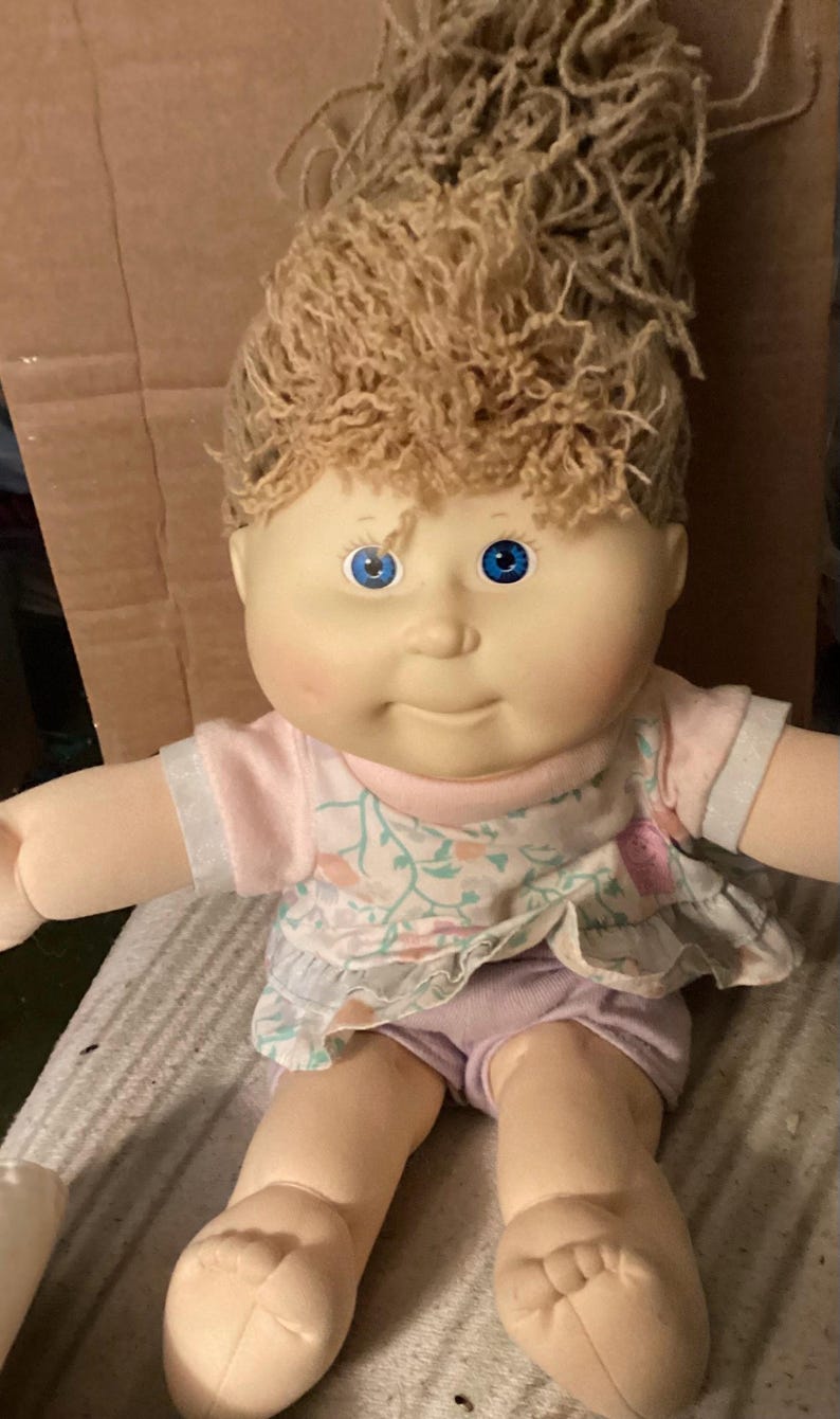 May include: A vintage doll with a soft, fabric body and a vinyl head. The doll has light skin, blue eyes, and light brown yarn hair. It wears a pink and floral print top and lilac shorts. The doll's arms and legs are extended.