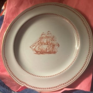 May include: White ceramic plate with a red ship illustration in the center. The plate has a red patterned border and a gold rim. The plate is likely vintage or antique.