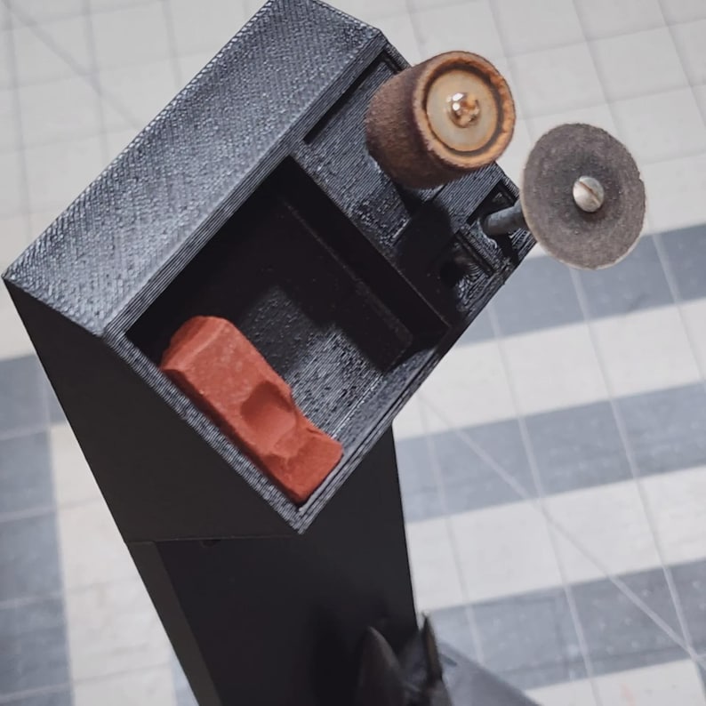 May include: Black 3D-printed tool holder with a compartment containing a red object. The holder has a section with two attached grinding wheels. The wheels are brown and gray.