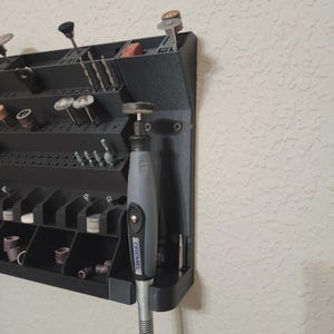 May include: A black, wall-mounted rotary tool organizer with various compartments and a Dremel rotary tool. The organizer holds bits, sanding drums, and other accessories. The tool is gray and black.