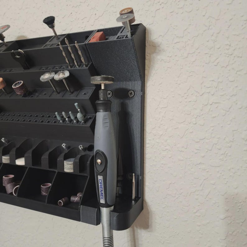 May include: A black, wall-mounted rotary tool organizer with various compartments and a Dremel rotary tool. The organizer holds bits, sanding drums, and other accessories. The tool is gray and black.