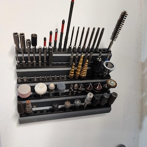 May include: A wall-mounted tool organizer with various drill bits, screwdriver bits, and other tools. The tools are arranged on a black metal rack, with a variety of sizes and shapes. Some tools have red accents.
