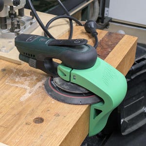 May include: A green and black power sander is clamped to a wooden workbench. The sander has a round sanding pad and a black handle. The workbench also has a router and a power cord.