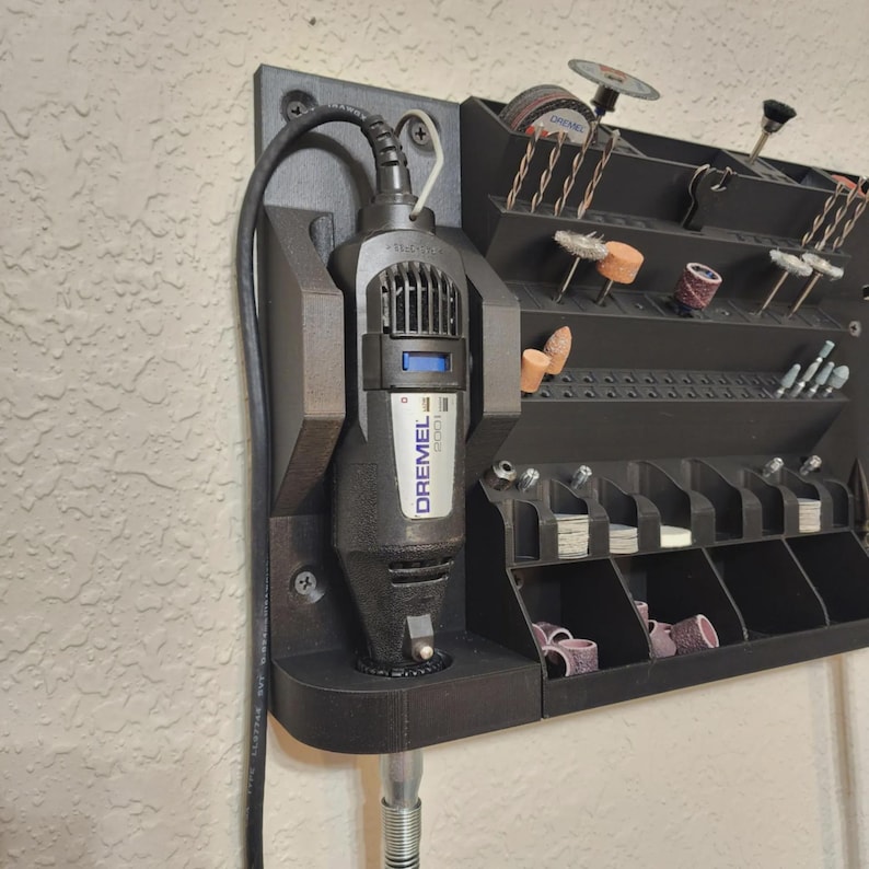 May include: A black Dremel rotary tool with the brand name visible, mounted on a black organizer. The organizer has multiple compartments and slots for various rotary tool accessories, including grinding wheels and sanding drums. The setup is attached to a textured wall.