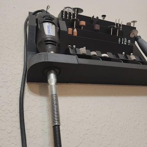 May include: A black Dremel rotary tool with a variety of bits and accessories organized in a tiered storage unit. The Dremel is mounted on the left side of the unit, with a flexible shaft extending downwards. The unit is mounted on a textured wall.