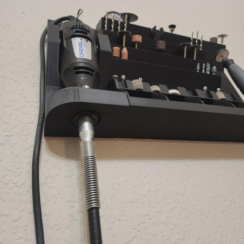 May include: A black Dremel rotary tool with a variety of bits and accessories organized in a tiered storage unit. The Dremel is mounted on the left side of the unit, with a flexible shaft extending downwards. The unit is mounted on a textured wall.