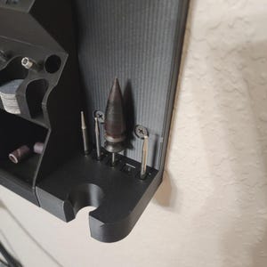 May include: A black, 3D-printed tool organizer mounted on a wall. It features various compartments and slots holding several metal drill bits and sanding drums. The organizer is designed to hold small tools and accessories, providing a neat storage solution.