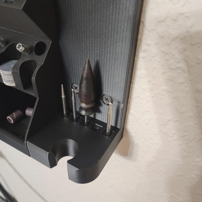 May include: A black, 3D-printed tool organizer mounted on a wall. It features various compartments and slots holding several metal drill bits and sanding drums. The organizer is designed to hold small tools and accessories, providing a neat storage solution.