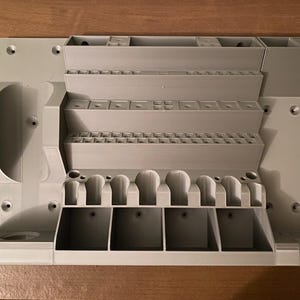 May include: A light gray, 3D-printed organizer with multiple compartments and slots. Features various sized storage areas, including small square slots, larger rectangular sections, and curved holders. The organizer is designed to hold tools or small items.