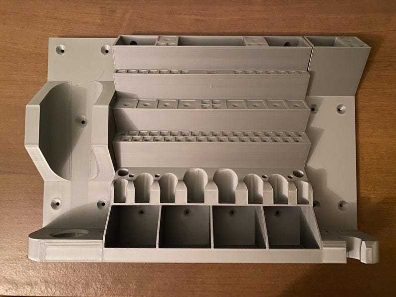 May include: A light gray, 3D-printed organizer with multiple compartments and slots. Features various sized storage areas, including small square slots, larger rectangular sections, and curved holders. The organizer is designed to hold tools or small items.