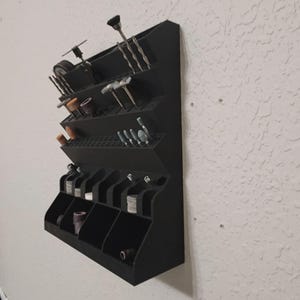 May include: A black wall-mounted organizer for small tools, featuring multiple angled shelves and compartments. Various tools, including drill bits and grinding accessories, are neatly arranged within the organizer, providing a structured storage solution.