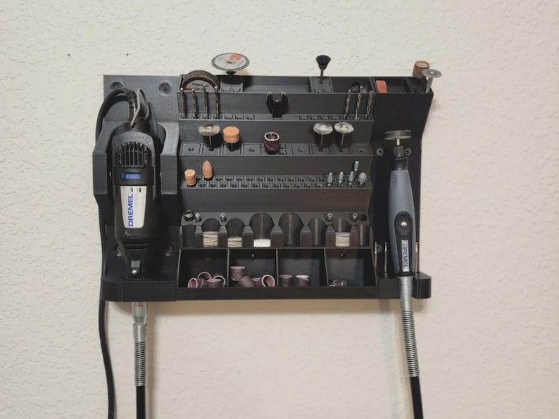 May include: A black wall-mounted organizer for rotary tools, featuring multiple compartments and holders for various bits and accessories. Two Dremel rotary tools are mounted on the organizer. The organizer is designed to keep tools and bits organized and accessible.