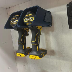May include: Two black and yellow Atomic drill drivers are mounted on a wall. The drills are secured in a dark blue holder. The drills have yellow accents and the brand name "ATOMIC" is visible on the top of the drills.