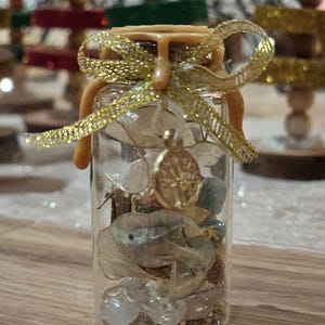 Peut inclure: Une petite bouteille en verre transparent remplie d'éléments décoratifs, notamment des fleurs séchées, de petites pierres et des paillettes dorées. La bouteille est scellée avec un bouchon marron clair et ornée d'un ruban doré et d'un petit charme doré. C'est un objet décoratif.