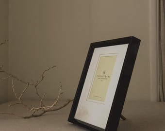 May include: A black rectangular picture frame with a white mat and a light yellow insert. The frame is propped up on a stand. A dried branch is in the foreground.