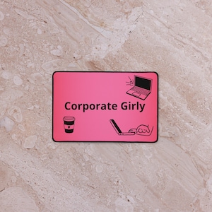 Puede incluir: Alfombrilla de ratón rectangular rosa con las palabras "Corporate Girly" en blanco. La alfombrilla presenta ilustraciones negras de un portátil, una taza de café y una persona usando un portátil.