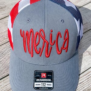 May include: A grey trucker hat featuring a red embroidered script text that reads 'Merica' on the front panel. The hat has a mesh back with a red, white, and blue pattern. A black rectangular sticker with white text reading 'RICHARDSON 112' is on the brim.