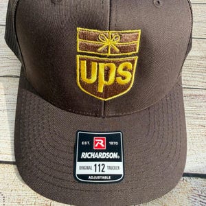 Op de afbeelding: Bruine truckerpet met een mesh achterkant en een gebogen klep. De voorkant toont het UPS-logo in goud en bruin. Een zwart-wit label onderaan luidt "RICHARDSON ORIGINAL 112 TRUCKER ADJUSTABLE."