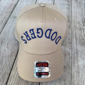 Op de afbeelding: Beige baseballpet met het woord "DODGERS" in blauw geborduurd. De pet heeft een gebogen klep en een stoffen label met de tekst "OTTO" en "CLASSIC FIT".