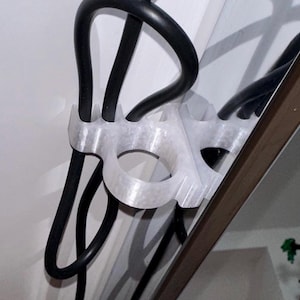 Peut inclure: Un organisateur de câbles blanc imprimé en 3D tenant des câbles noirs contre un mur blanc. L'organisateur comporte plusieurs boucles et une ouverture circulaire pour fixer les câbles. La conception est simple et fonctionnelle.