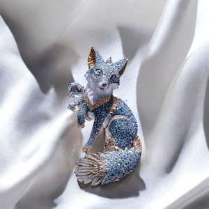 May include: A blue jeweled fox brooch with a white and yellow chest and a long, feathery tail. The brooch is set against a white satin background.